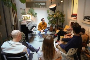 singing teacher delivers a singing lesson to a group of students sat around piano in a modern studio with plants and lighting