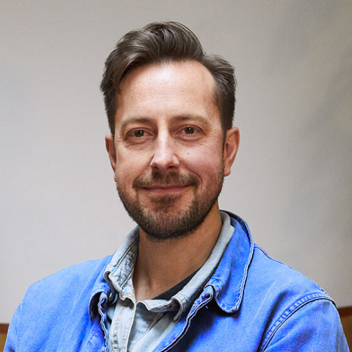 A professional headshot of a smiling man with short brown hair and a beard, wearing a blue jacket over a denim shirt.