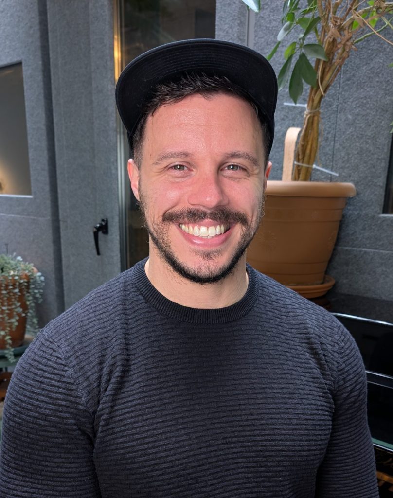 Mike Dzyngel, vocal coach at City Vocal Coach, smiling in front of a piano and potted plants